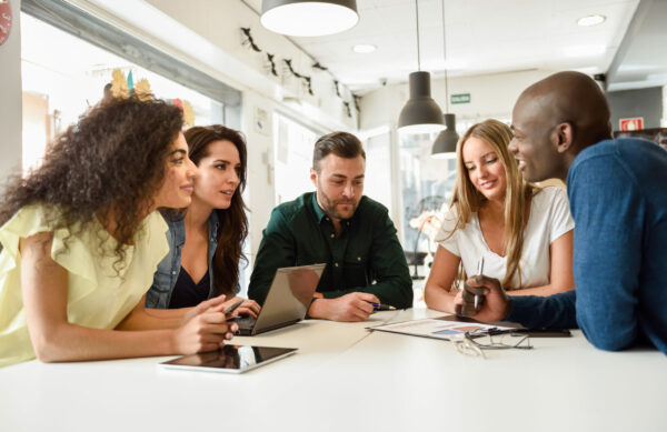 Five young people studying with laptop and tablet computers on white desk. Beautiful girls and guys working toghether wearing casual clothes. Multi-ethnic group smiling.